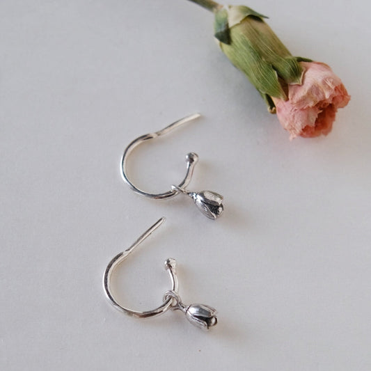 A pair of silver-colored earrings shaped like tulips, displayed next to a withered pink carnation on a white surface.