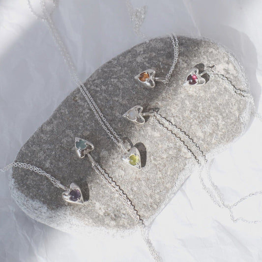 A silver pendant in the shape of a heart, adorned with multiple colored gemstones, displayed on a rock surface with a chain attached.